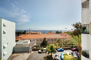 A multi-story building with a red-tiled roof, a parking area with several cars, and a view of the sea in the background.