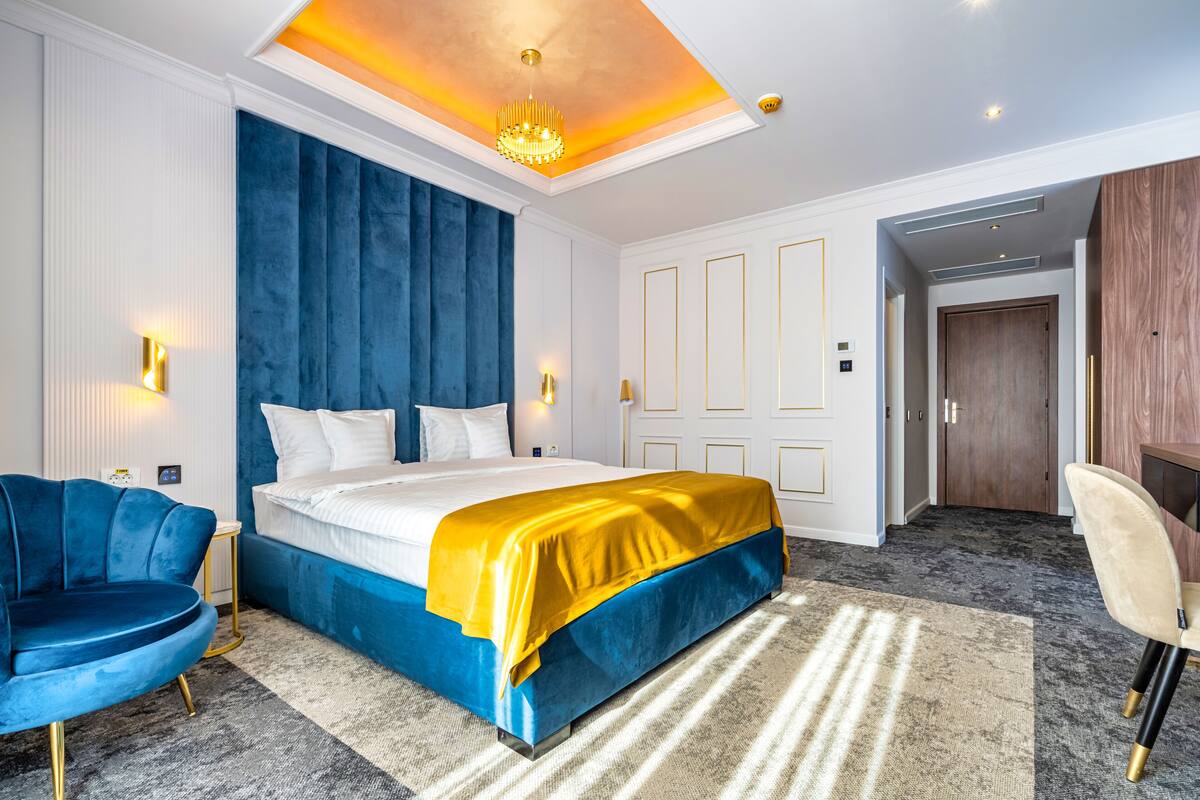 A modern hotel room with a large bed, a blue accent wall, a yellow bedspread, a blue armchair, and a wooden wardrobe.