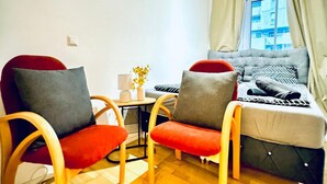 Une petite pièce avec un canapé, deux fauteuils, une petite table avec une lampe et une fenêtre avec des rideaux.