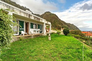 A white building with green shutters, a grassy lawn, and a mountain in the background. - Holiday Home 'Blissful Haven - Soulful Stays' with Sea View, Private Terrace and Wi-Fi (Porto Moniz)