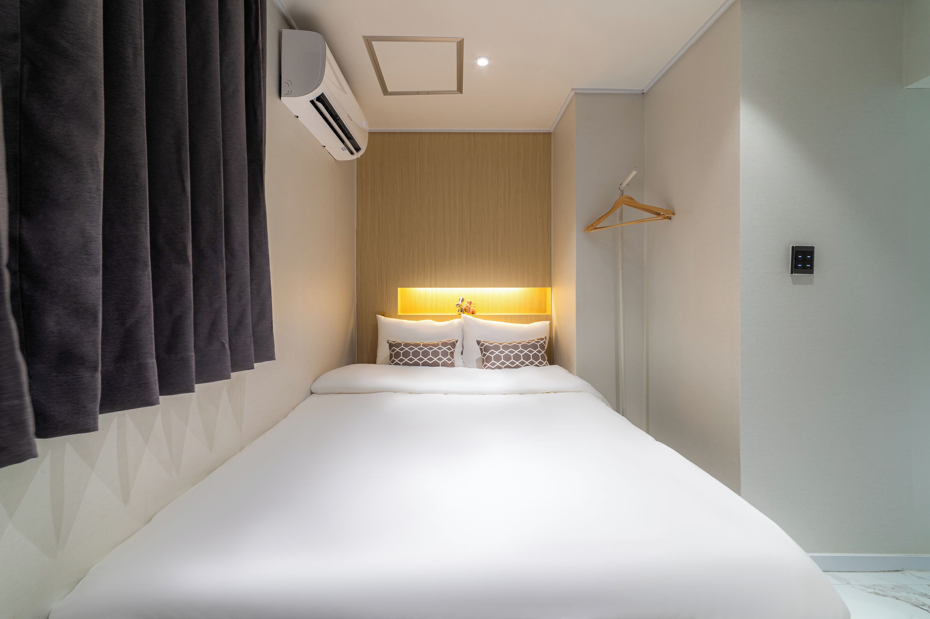 A modern hotel room with a large bed, a bedside table with a lamp, and a wall-mounted air conditioner.