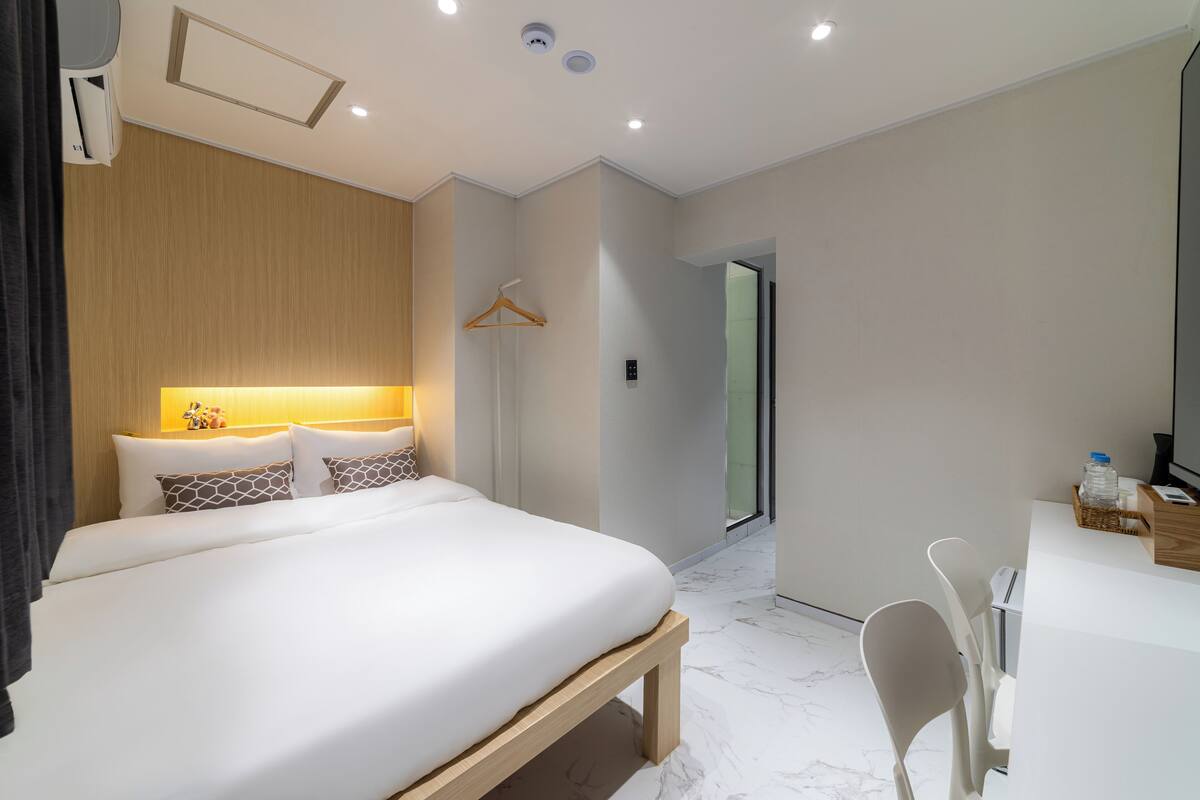 A modern hotel room with a large bed, bedside table, and a desk with a chair.