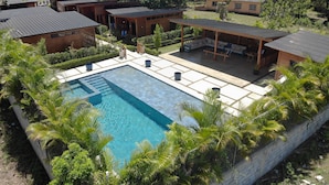 A pool area with a clear blue pool, surrounded by a stone wall and lush greenery. There are several wooden buildings with metal roofs, and a covered seating area with outdoor furniture. - Villa Dulce Refugio (Cabrera)