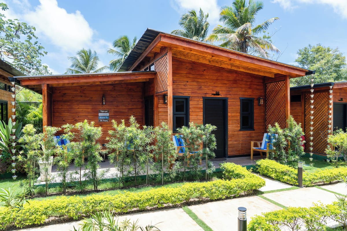 Una cabaña de madera con porche, rodeada de jardines cuidados y palmeras.