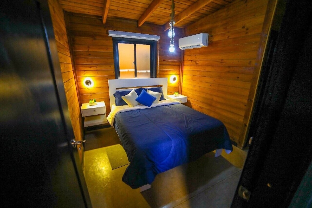 Una habitación pequeña de madera con una cama, mesitas de noche y una ventana.