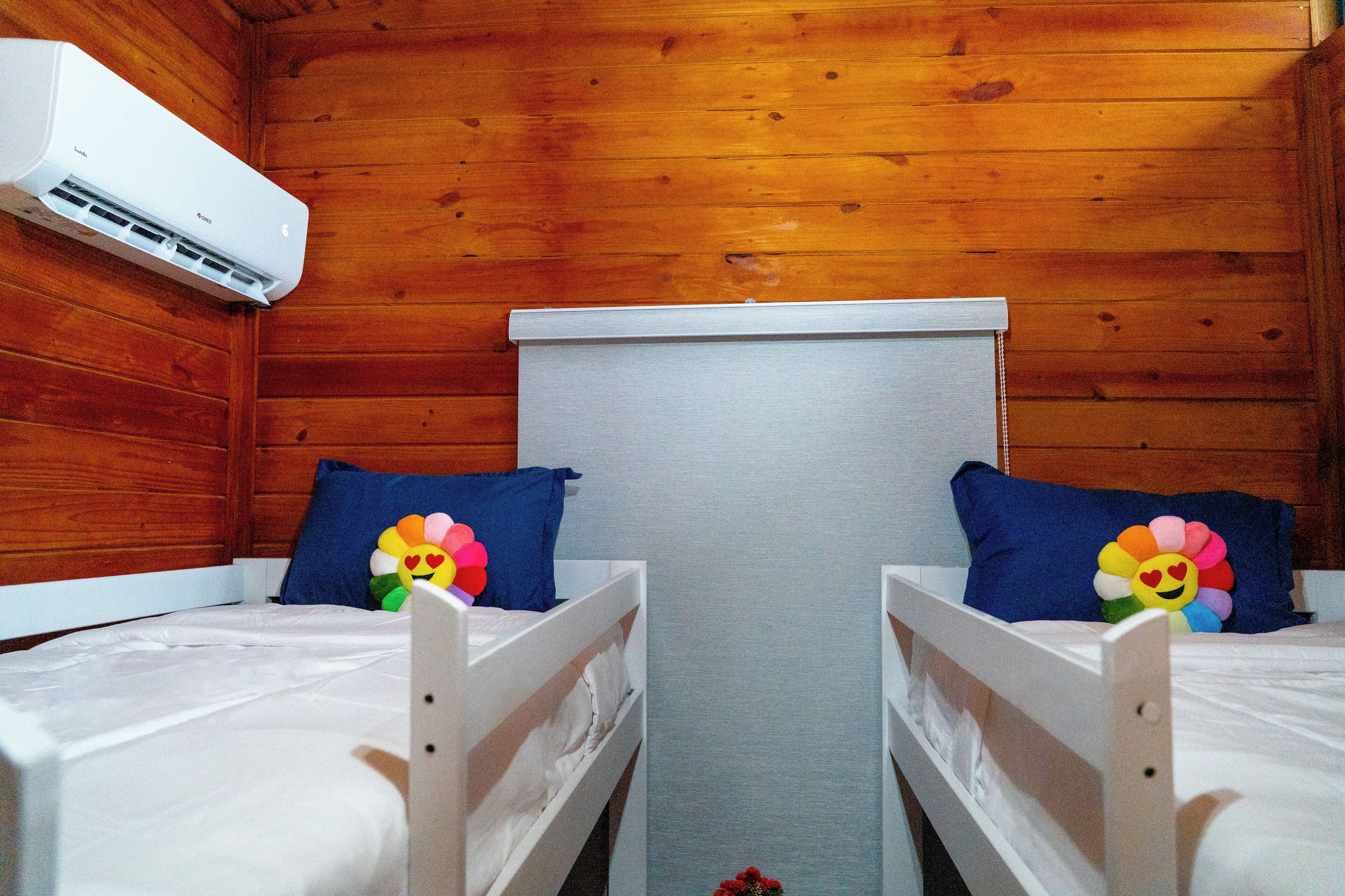 A room with two single beds, wooden walls, a white air conditioning unit, and a window with a white blind.