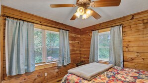 A wooden cabin bedroom with a bed, a ceiling fan, and two windows with curtains.