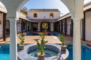 A pool area with a tiled hot tub, potted plants, and a building with arched doorways in the background.