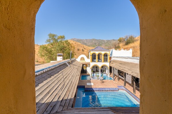 A pool area with a wooden deck, a small building with arches, and a view of hills.