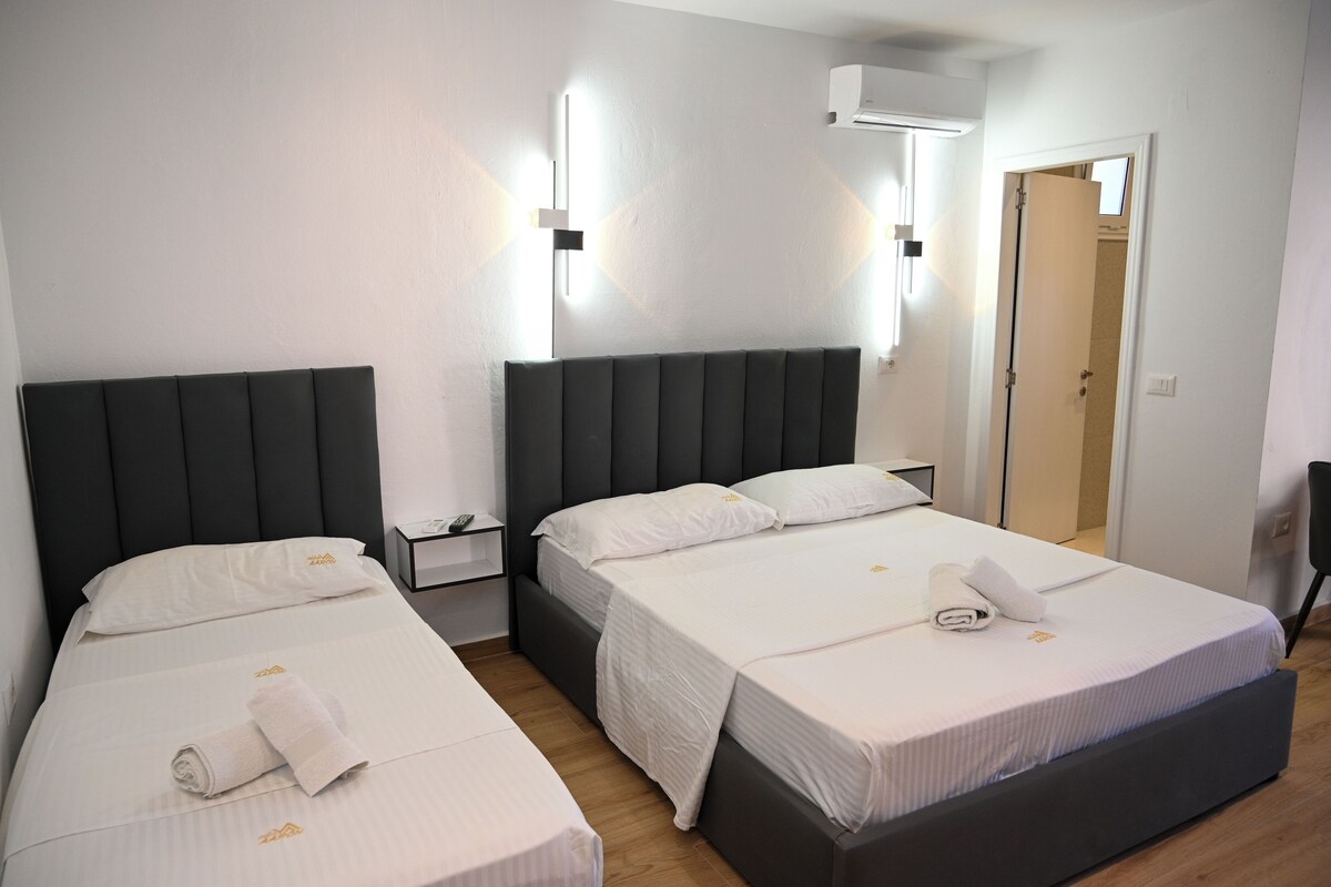 Habitación de hotel con dos camas, cada una con cabecero oscuro y ropa de cama blanca. Hay mesitas de noche con lámparas, una lámpara de pared y una puerta que da a otra habitación.