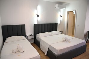 Habitación de hotel con dos camas, cada una con cabecero oscuro y ropa de cama blanca. Hay mesitas de noche con lámparas, una lámpara de pared y una puerta que da a otra habitación.