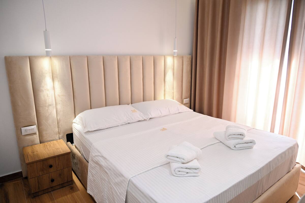 A neatly made bed with white linens and two folded towels, a wooden nightstand with a drawer, and a modern headboard.