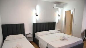 A hotel room with two beds, each with a dark headboard and white bedding. There are bedside tables with lamps, a wall-mounted light fixture, and a door leading to another room. - Hotel Vila Aaron (Manëz)