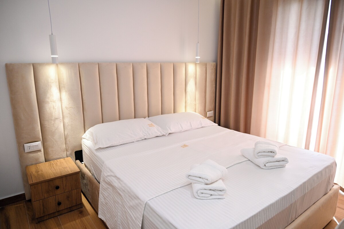 A neatly made bed with white linens and two folded towels, a wooden nightstand with a drawer, and a modern headboard.
