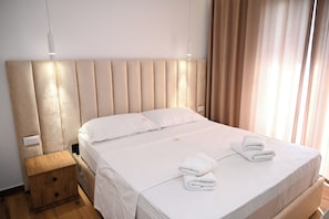 A neatly made bed with white linens and two folded towels, a wooden nightstand with a drawer, and a modern headboard. - Hotel Vila Aaron (Durrës)