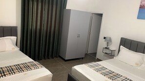 Habitación de hotel con dos camas, un armario, una mesita de noche con lámpara y una cortina.