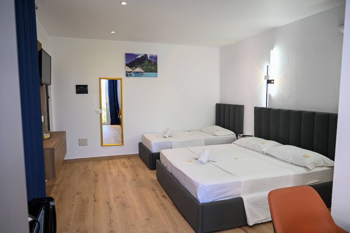 A hotel room with two beds, a wooden floor, a flat-screen TV, and a painting of a tropical scene.