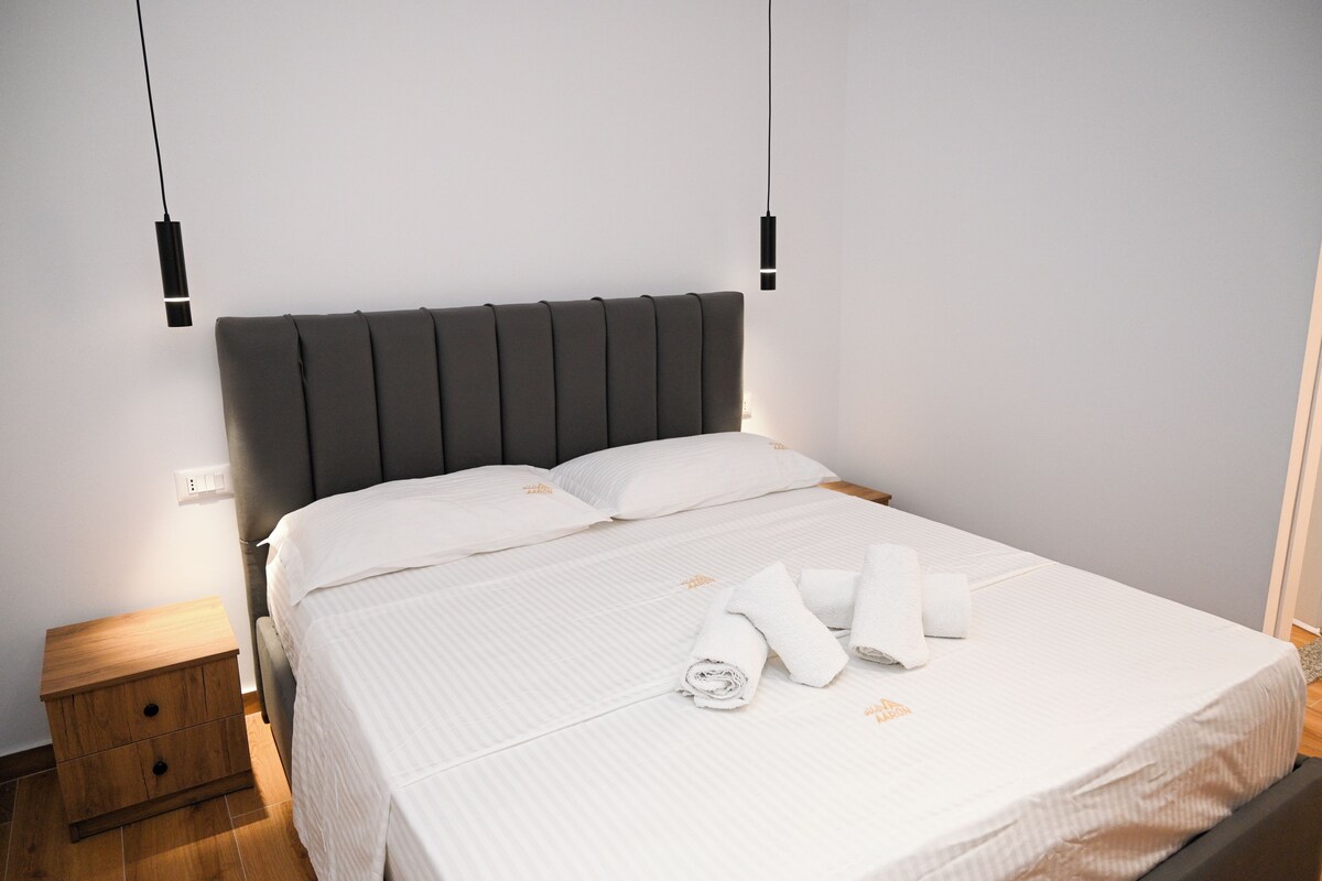 A neatly made bed with white linens and rolled towels, a wooden nightstand, and a modern headboard.