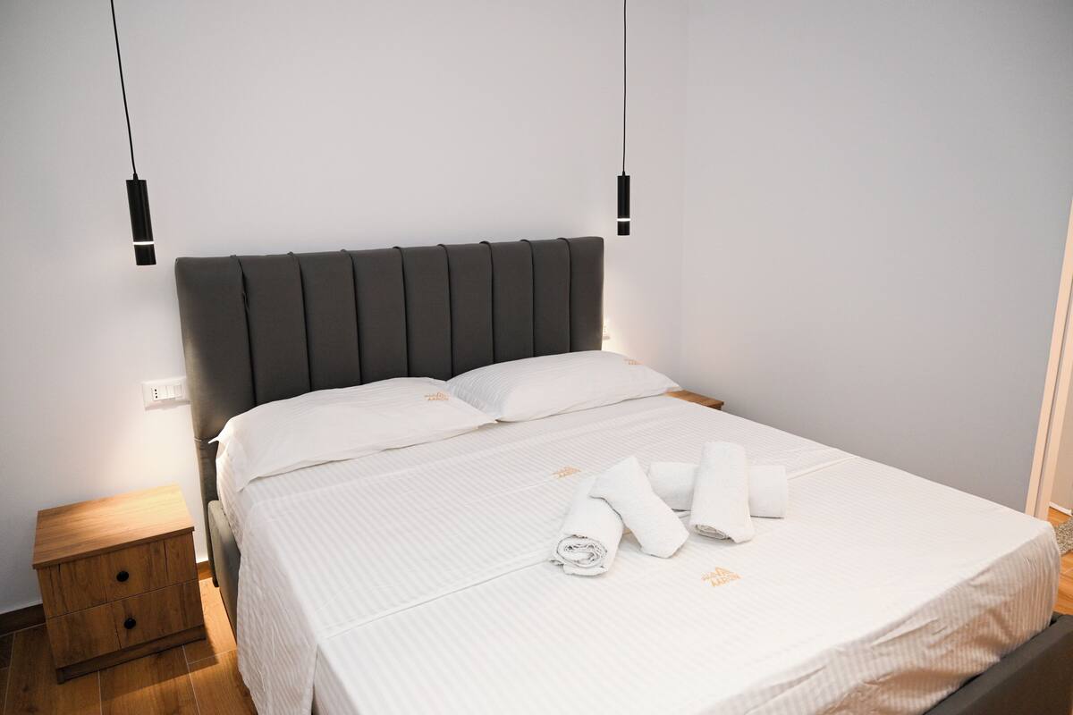 A neatly made bed with white linens and rolled towels, a wooden nightstand, and a modern headboard.