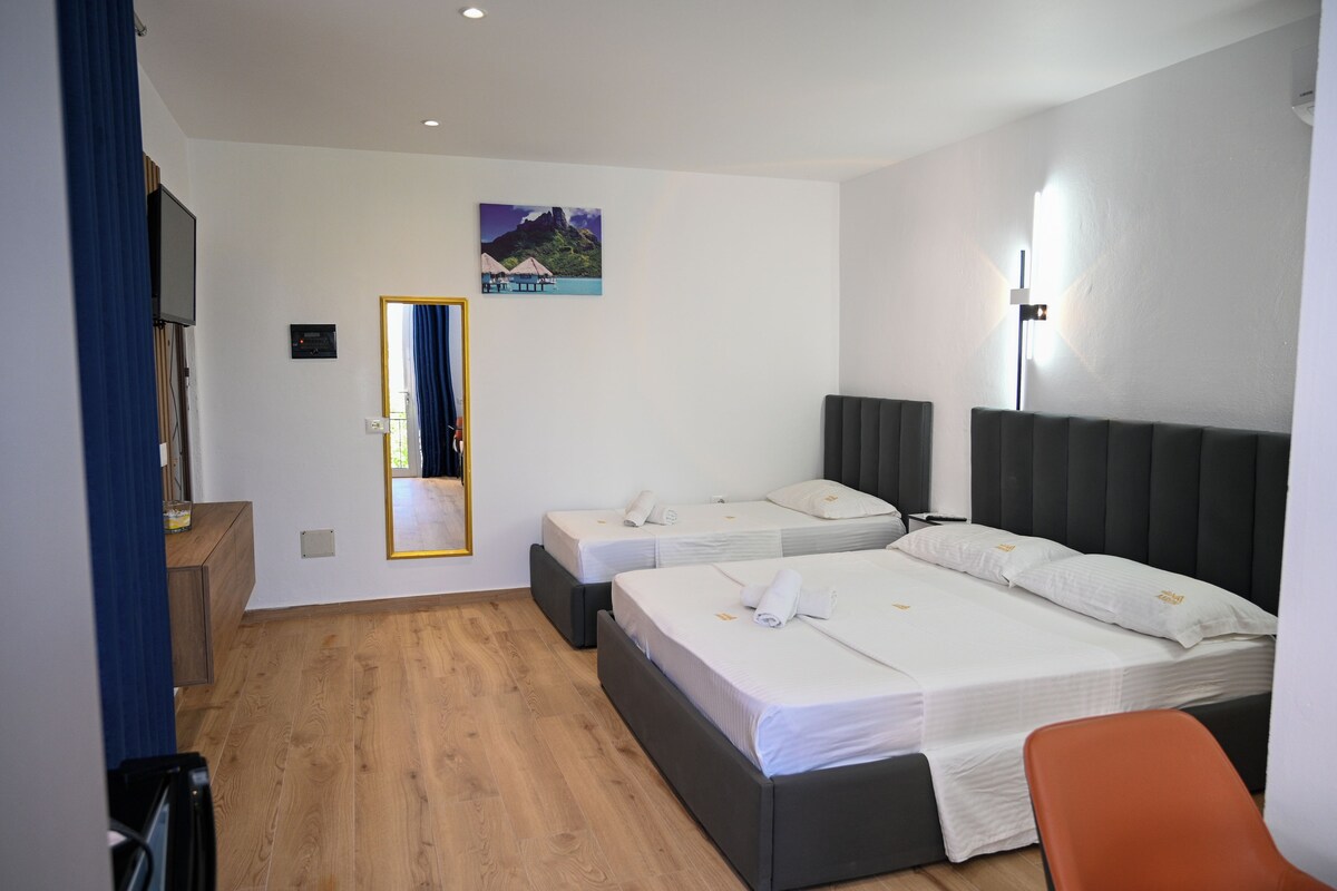 A hotel room with two beds, a wooden floor, a flat-screen TV, and a painting of a tropical scene.