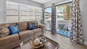 A living room with a beige sofa, decorative pillows, a coffee table with a fruit bowl, and a view of a pool through a glass door. - Miramar Beach Oasis - 4ppl (Miramar Beach)