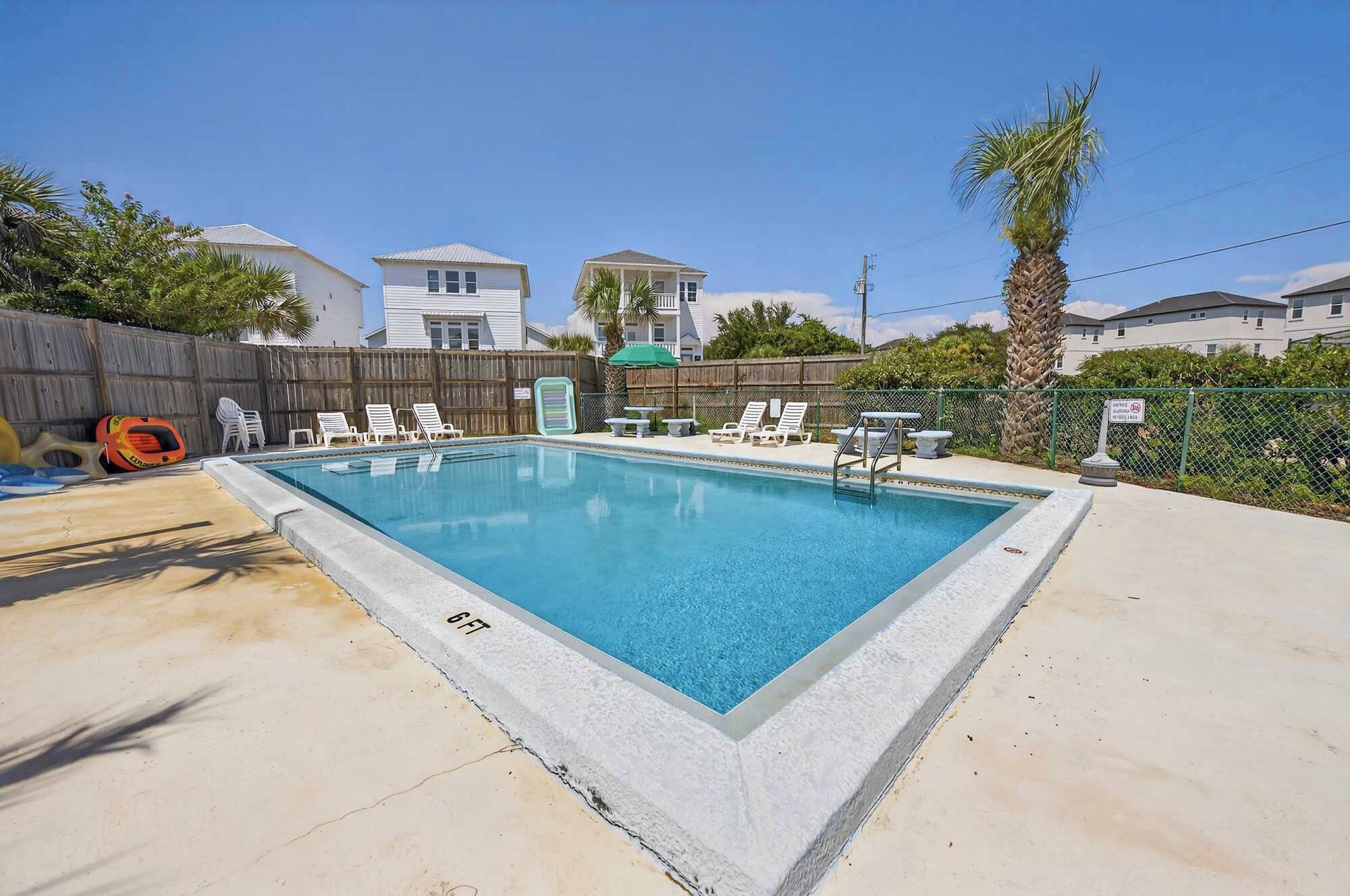 A rectangular swimming pool with a concrete edge, surrounded by a wooden fence, and equipped with lounge chairs and a table.