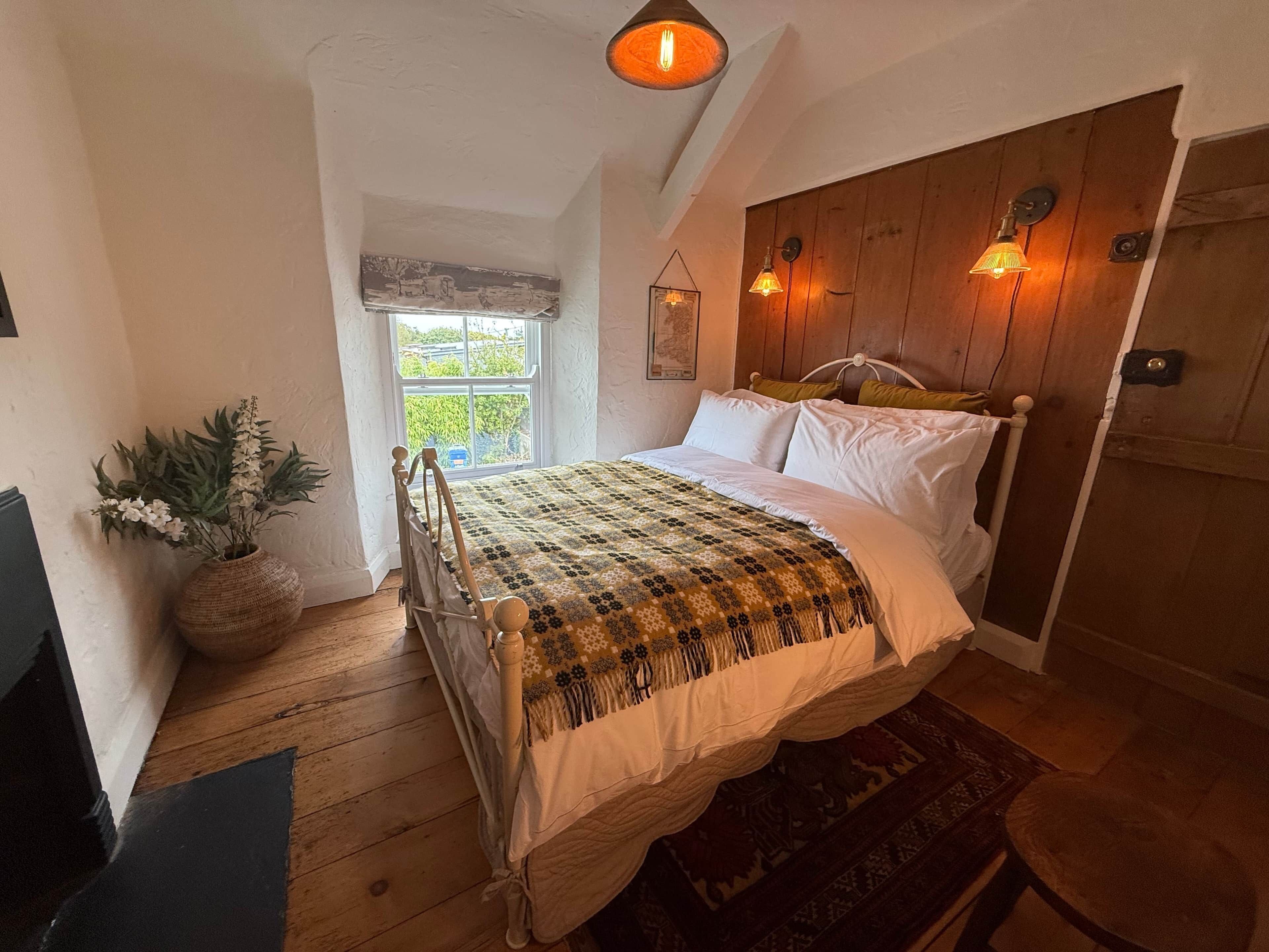 A bedroom with a wooden bed, a window, a potted plant, and a wall-mounted light fixture.
