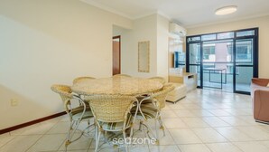 A dining area with a round table and wicker chairs, a sofa, and a large window with a view. - Apt w Balcony nd Barbecue in Seaside Condom Uar301 (Itapema)