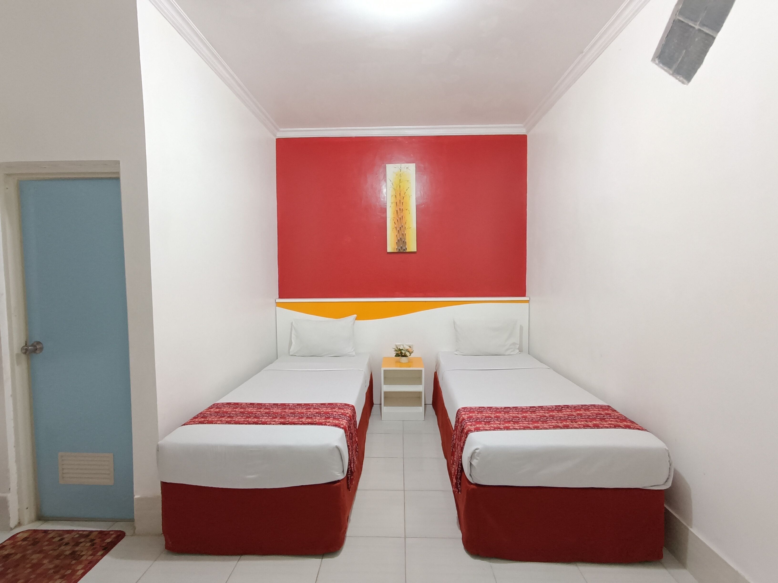 Quarto pequeno e colorido com duas camas simples, uma mesinha com uma planta e uma porta à esquerda.