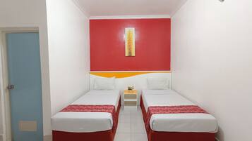 Quarto pequeno e colorido com duas camas simples, uma mesinha com uma planta e uma porta à esquerda.
