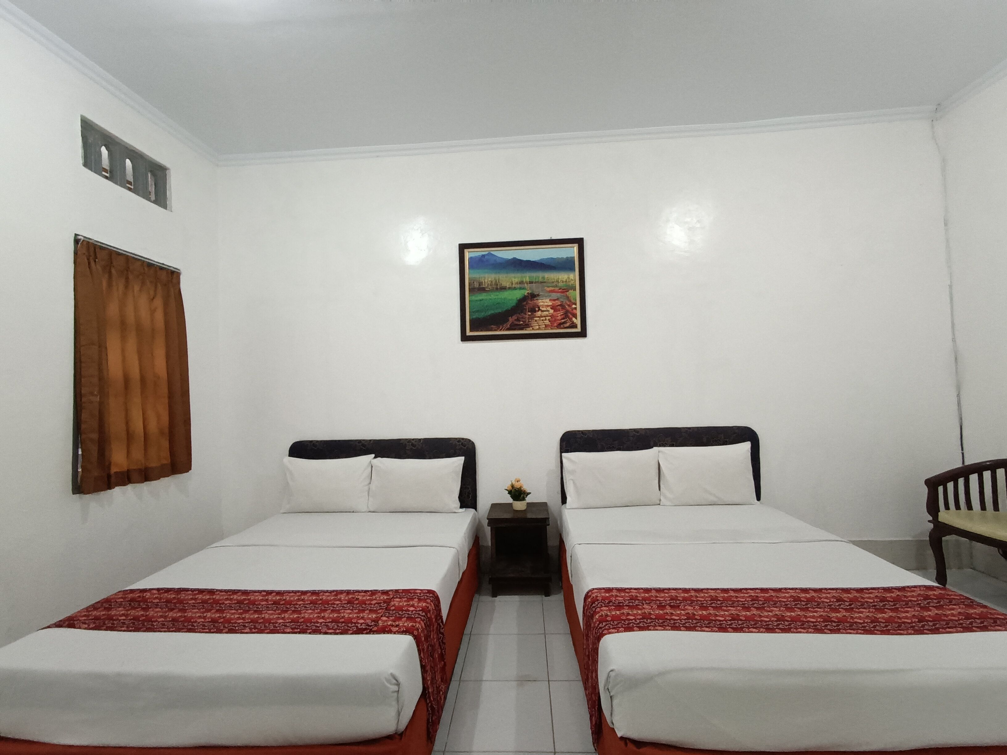 A room with two beds, a small table with a vase, and a painting on the wall.