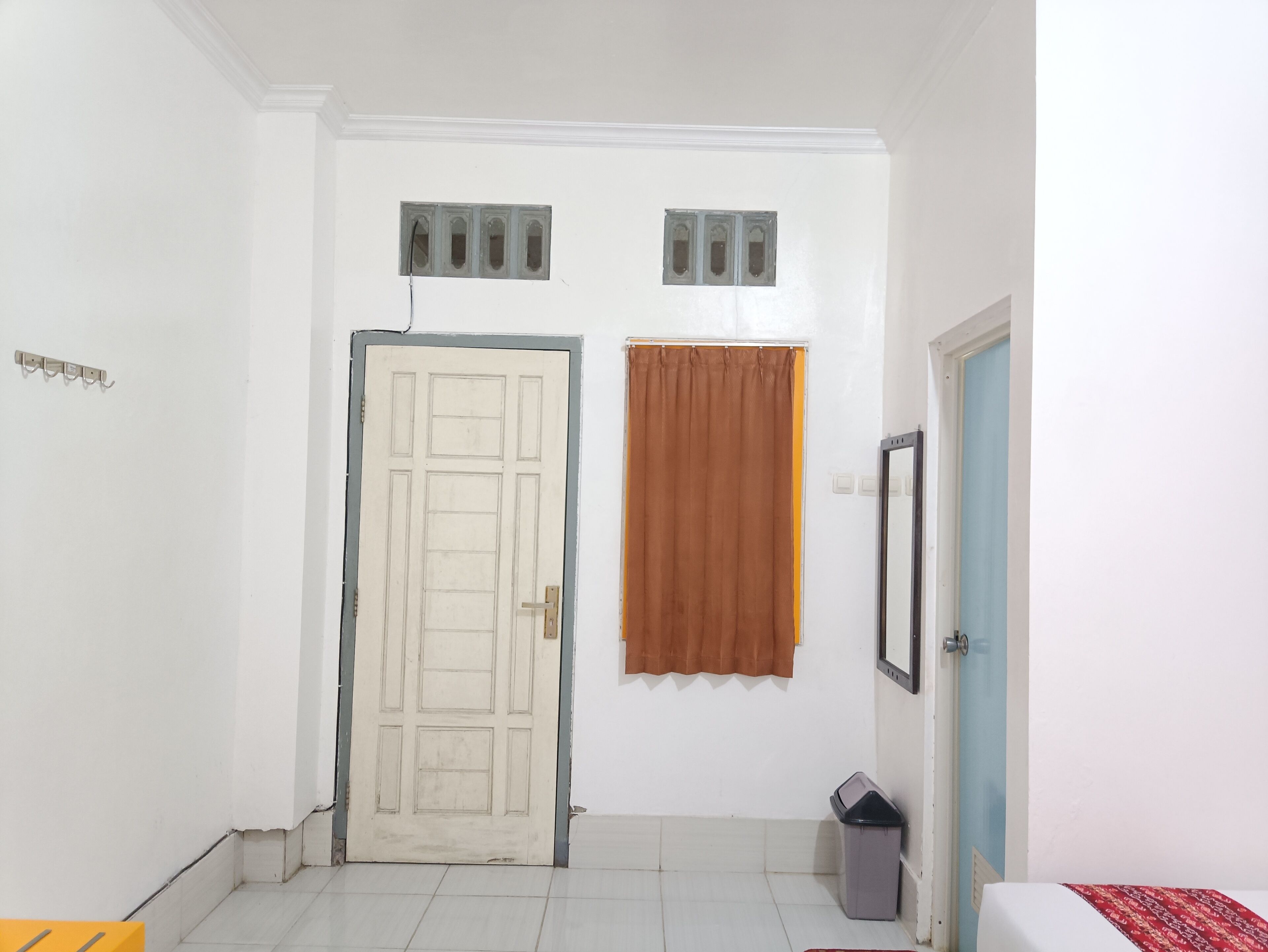 A hallway with a white door, a brown curtain, a trash bin, and a bed with a red patterned bedspread.