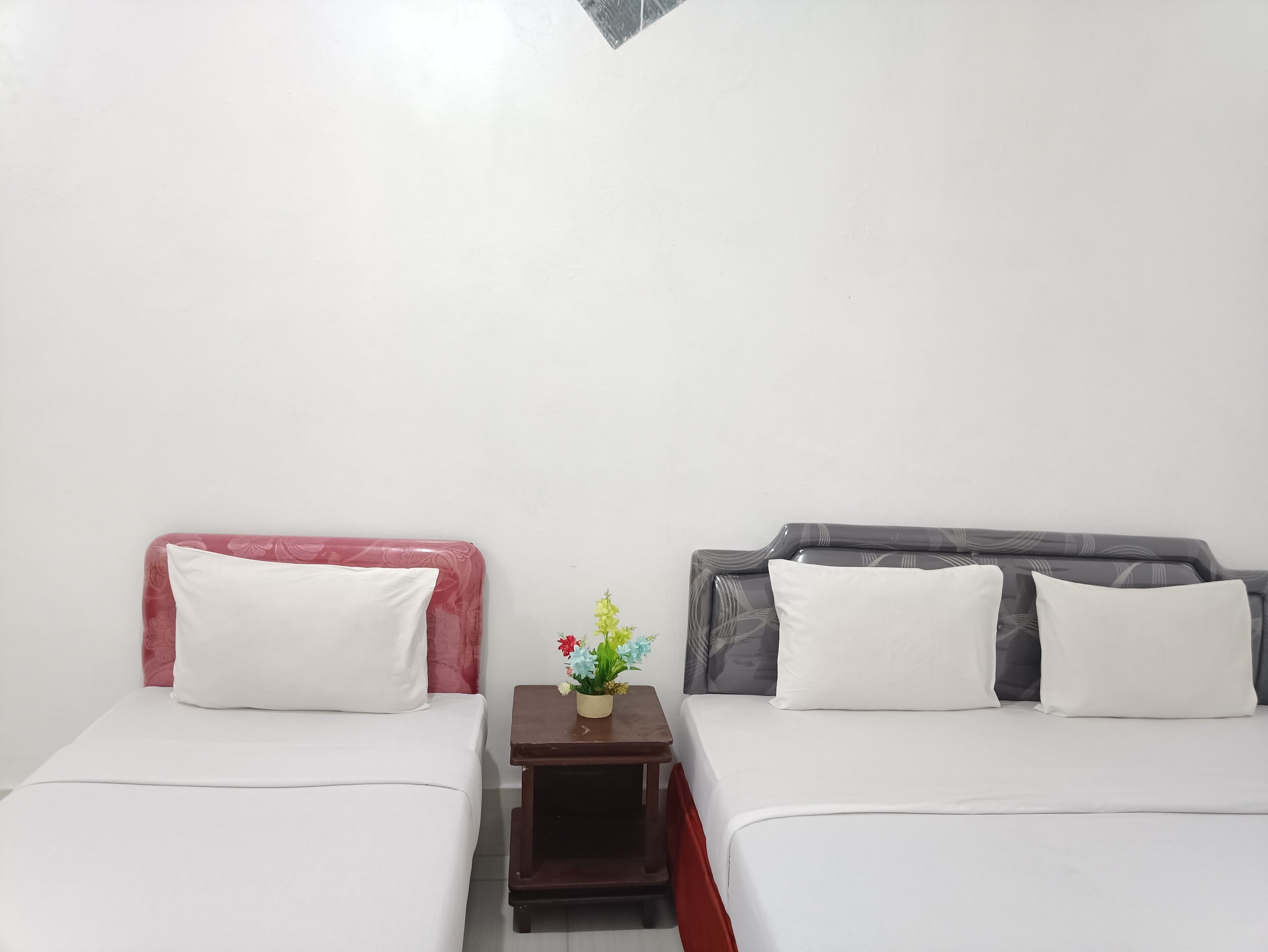 Two single beds with white bedding, a small wooden nightstand with a vase of flowers, and a plain white wall.