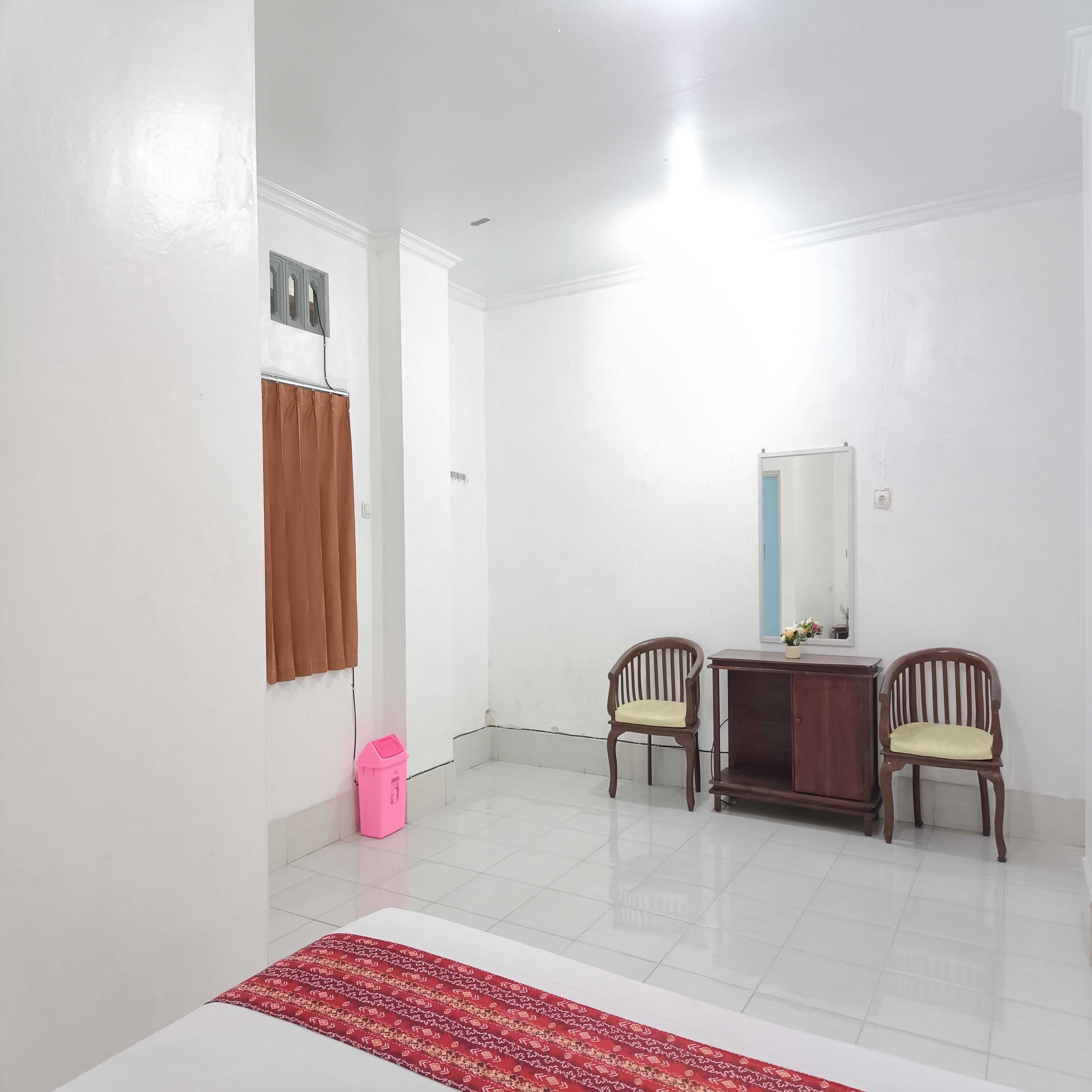 A room with white walls, a wooden cabinet, two wooden chairs, a small table, a mirror, and a red patterned rug.