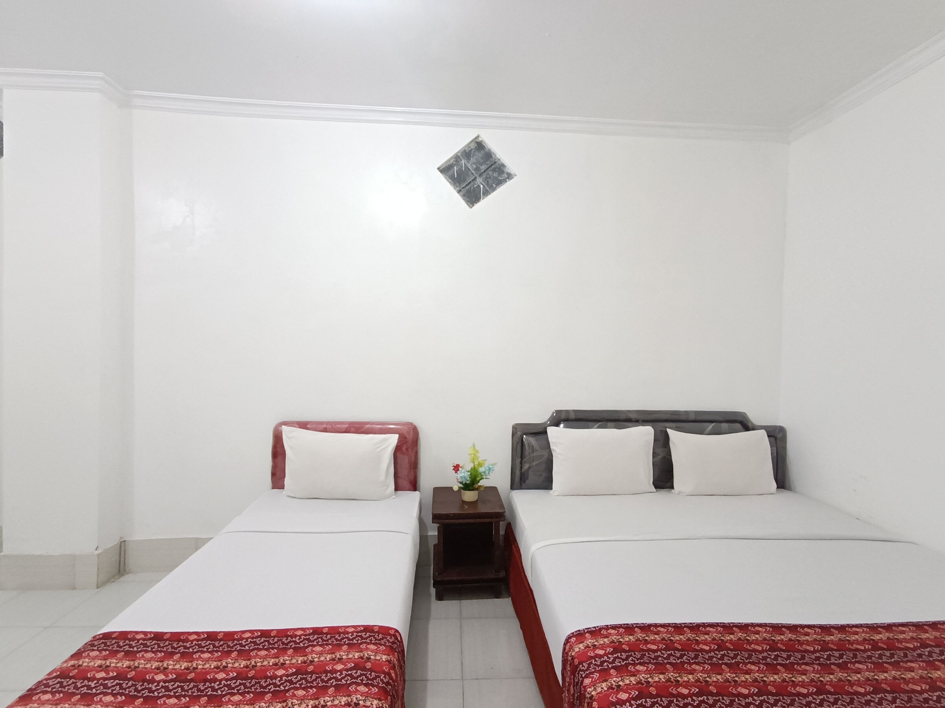 Habitación de hotel con dos camas, cada una con una colcha roja estampada y almohadas blancas. Entre las camas hay una mesita de noche de madera con una planta en maceta. Las paredes son blancas y hay un pequeño adorno colgando en la pared.