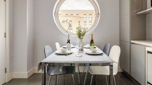 A dining area with a round window, a table set for two, and a view of buildings outside. - The Tower Bridge Stateroom — 4‑Bed Penthouse w/ AC (London)