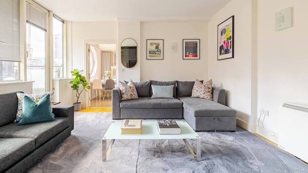 A modern living room with a grey sectional sofa, a smaller grey sofa, and a coffee table. There are two framed pictures on the wall, a potted plant, and a window with blinds. - The Tower Bridge Stateroom — 4‑Bed Penthouse w/ AC (London)