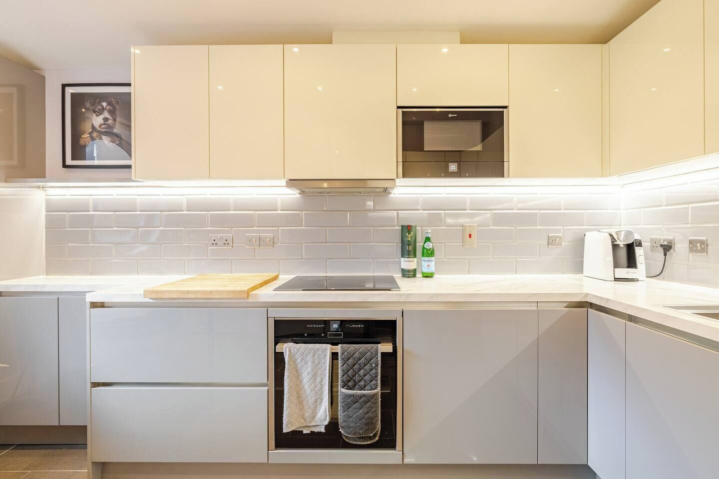 A modern kitchen with white cabinetry, a built-in oven, a countertop with a cutting board, and a coffee machine.