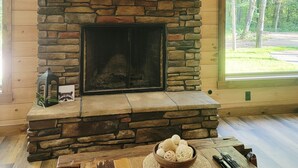 A stone fireplace with a flat-screen TV above it, a wooden table with a bowl of balls, and a window with a view of trees. - Cozy 2-bedroom cabin in near Gambier, peaceful woodland setting (Gambier)