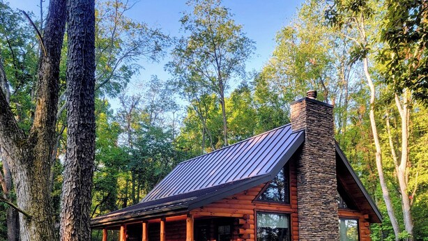 A log cabin with a stone chimney, large windows, and a porch. - Cozy 2-bedroom cabin in near Gambier, peaceful woodland setting (Gambier)