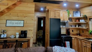 A cozy cabin interior with a wooden staircase, a living area with a sofa, a kitchen with a refrigerator, and a dining table. - Cozy 2-bedroom cabin in near Gambier, peaceful woodland setting (Gambier)