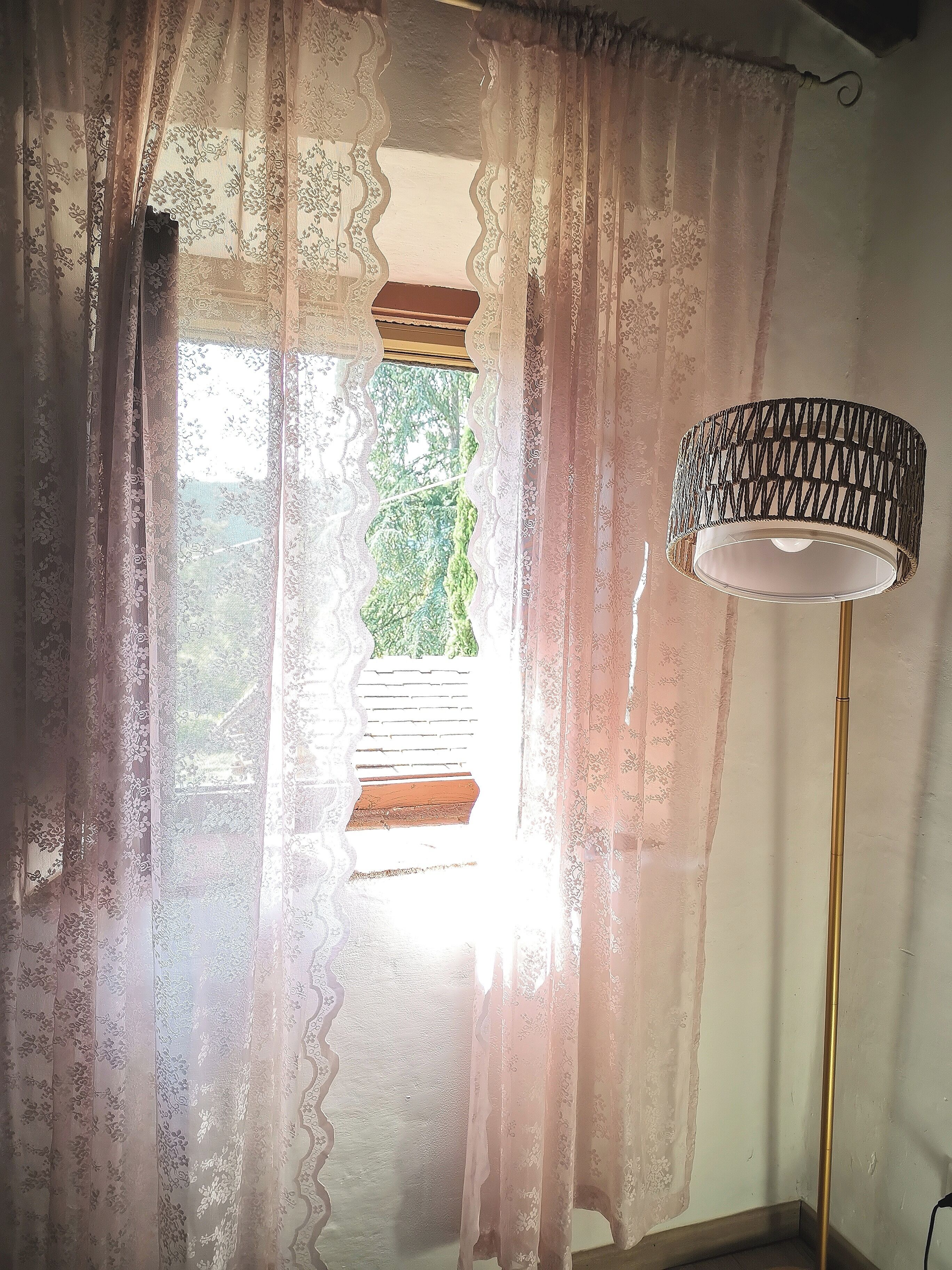 Ein Zimmer mit leichtem Vorhang, einem Fenster mit Blick auf Grünflächen und einer Stehlampe.