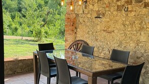 An outdoor patio with a glass-top table and wicker chairs, set against a stone wall with a view of a green landscape.