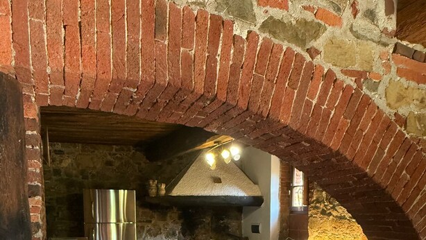 A rustic kitchen with a brick archway, wooden flooring, a stainless steel refrigerator, and a central island with stools.