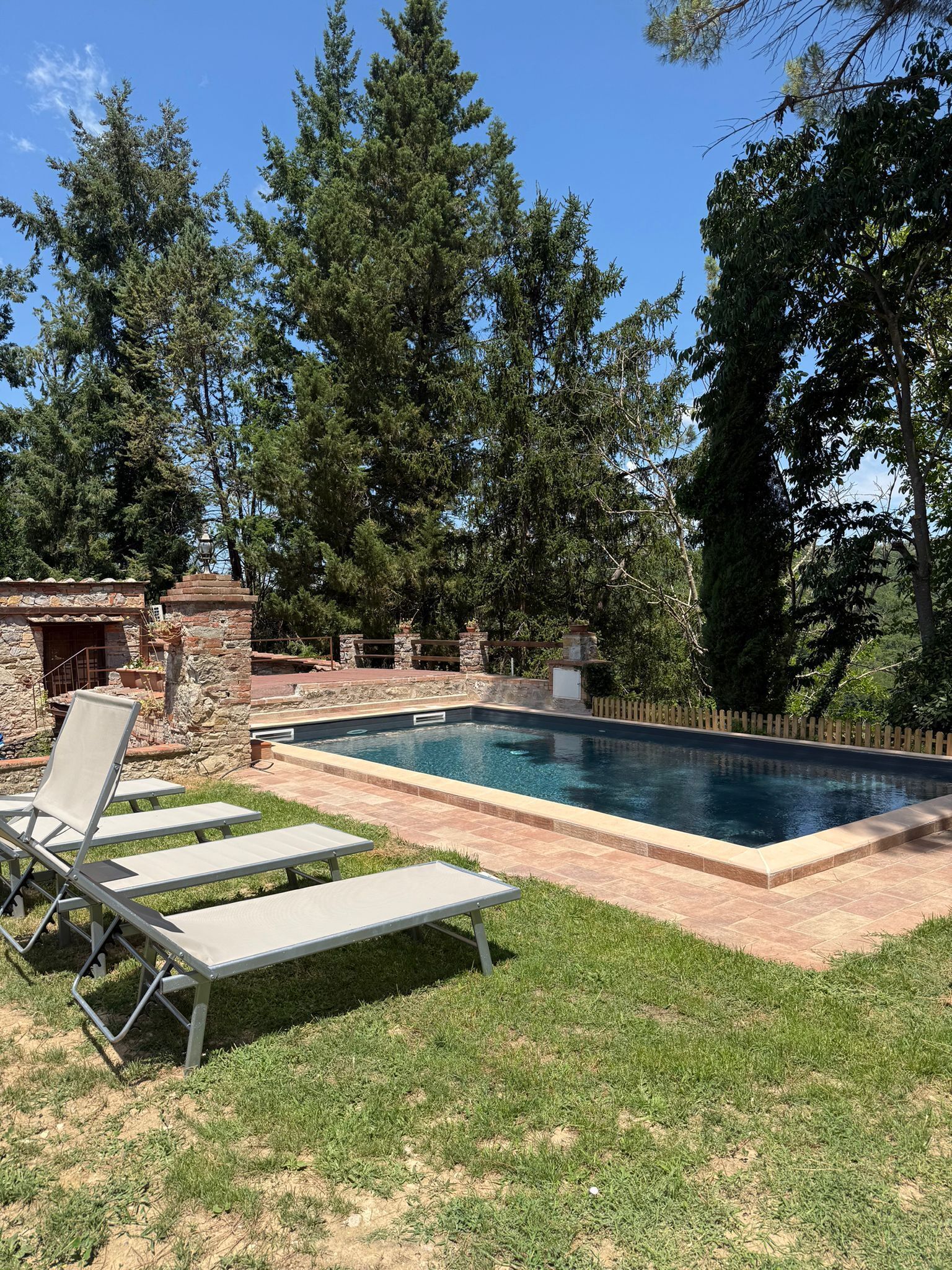 A swimming pool with lounge chairs, surrounded by a stone patio and a wooden fence.