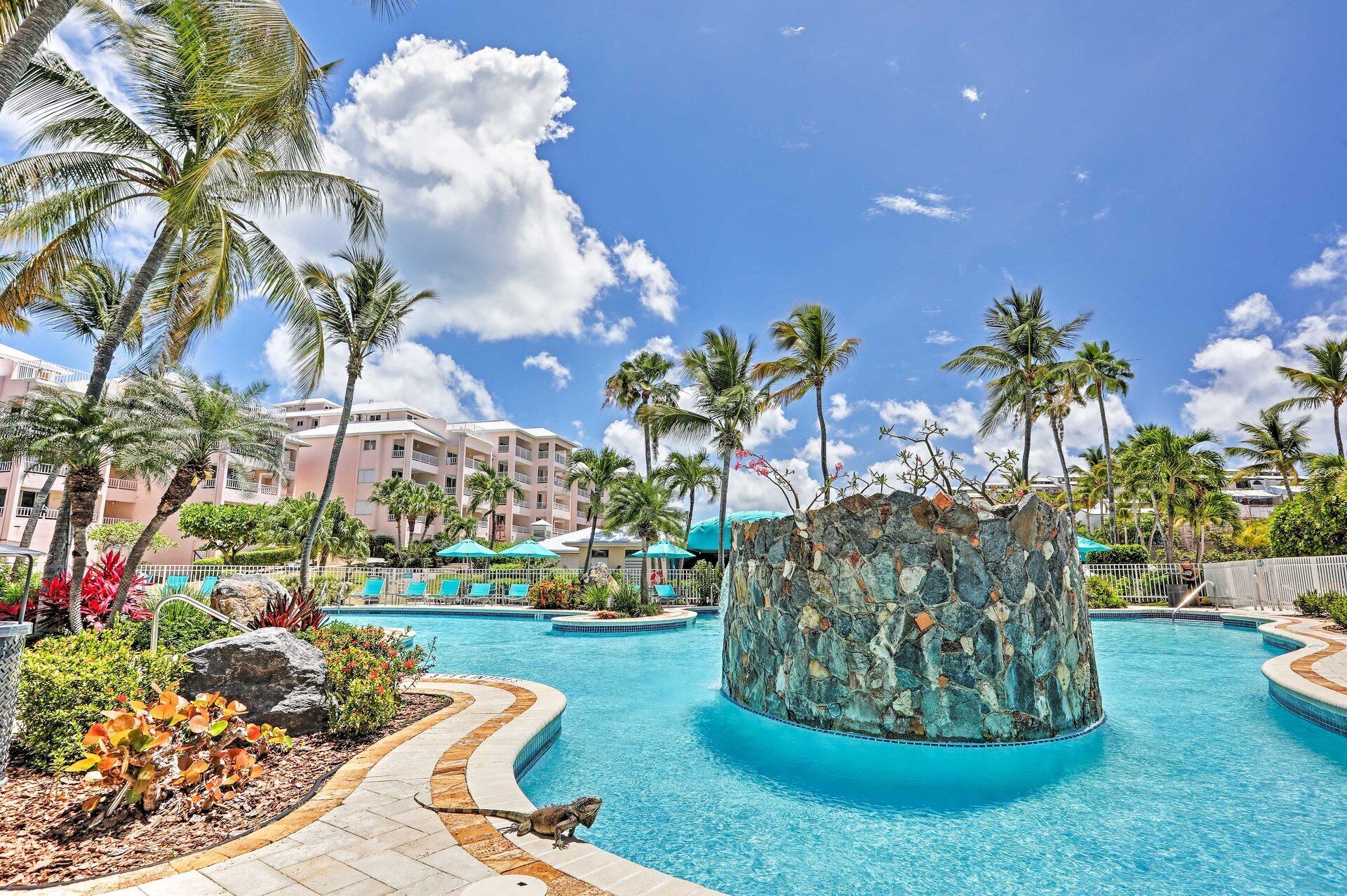 A resort pool area with palm trees, a stone water feature, and a building with balconies in the background.