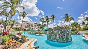 A resort pool area with palm trees, a stone water feature, and a building with balconies in the background.