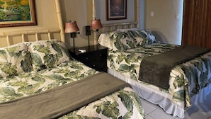 A bedroom with two beds, each with a tropical leaf-patterned bedspread, a bedside table with lamps, and a framed painting on the wall.
