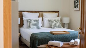 A neatly made bed with a green blanket, two pillows with a striped pattern, a bedside table with a lamp, and a tray with towels. - Domaine Robert (Rognes)
