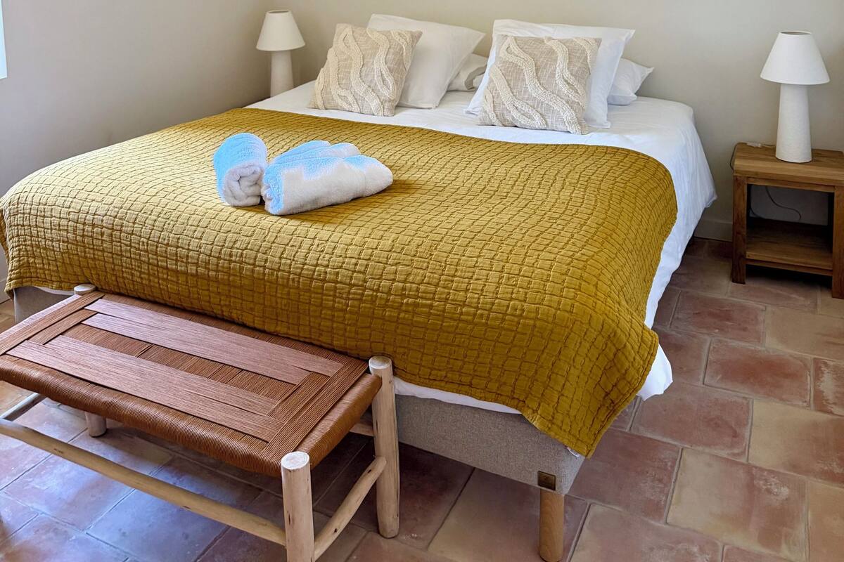 A neatly made bed with a yellow quilt, white pillows, and a wooden bench at the foot of the bed.